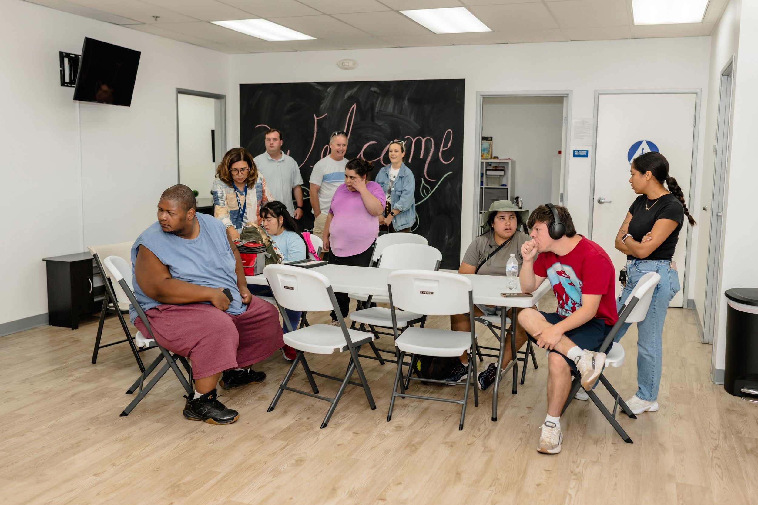 A diverse group of people gathered in a room with white walls and a black chalkboard that says 'Welcome.' Some are sitting at tables, others are standing, and one woman is wearing headphones. The group appears to be participating in a workshop or meeting.