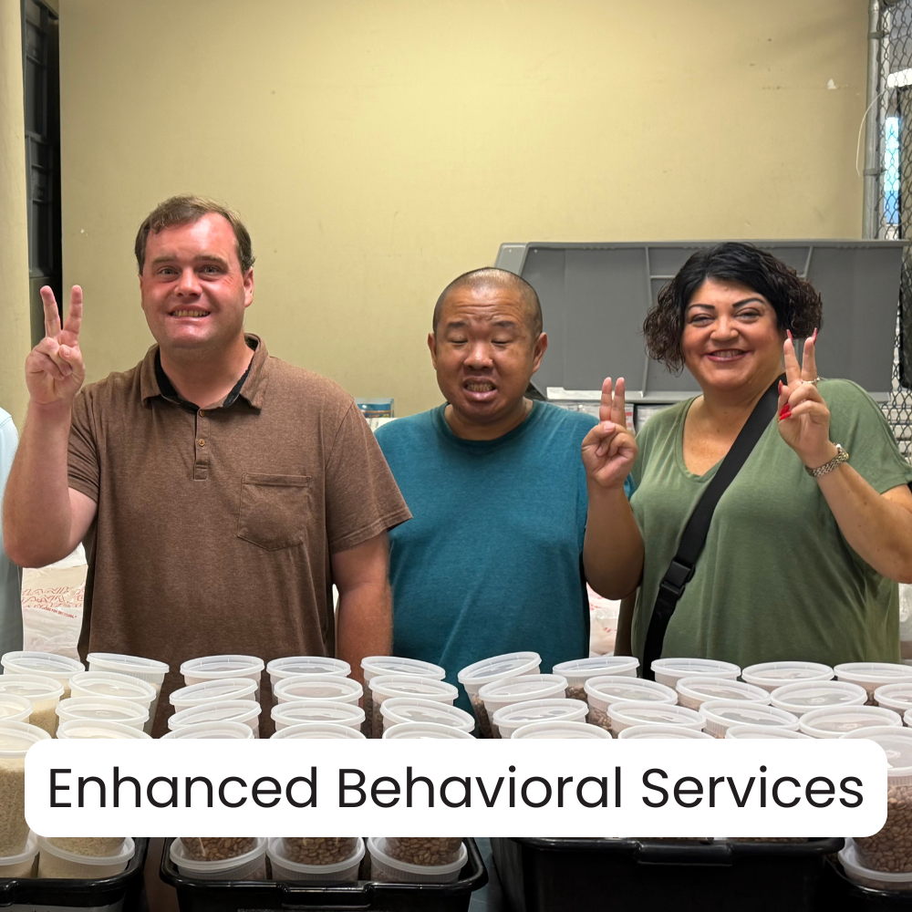 Three people standing behind tables filled with containers of beans, smiling and making peace signs. The setting appears to be a community center or similar indoor space.