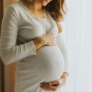 A pregnant woman standing near a window, holding her belly with both hands.