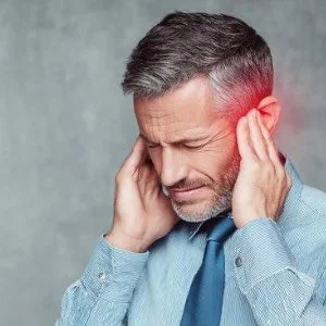 A middle-aged man holding his head with both hands, appearing to have a headache or ear pain, with a pained expression on his face.
