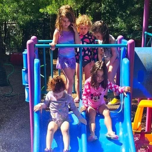 Six young girls playing on a colorful playground slide outdoors in sunny weather.