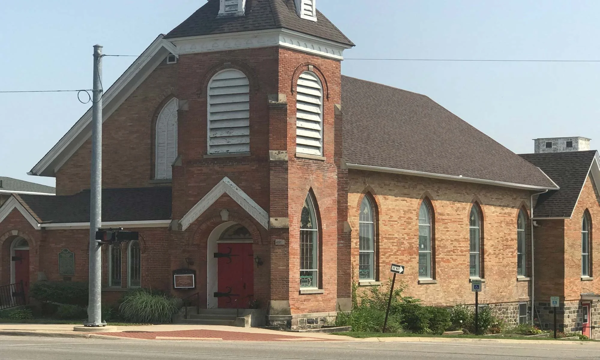 A brick church with arched windows and a steeple, located along a street with a utility pole and street signs.