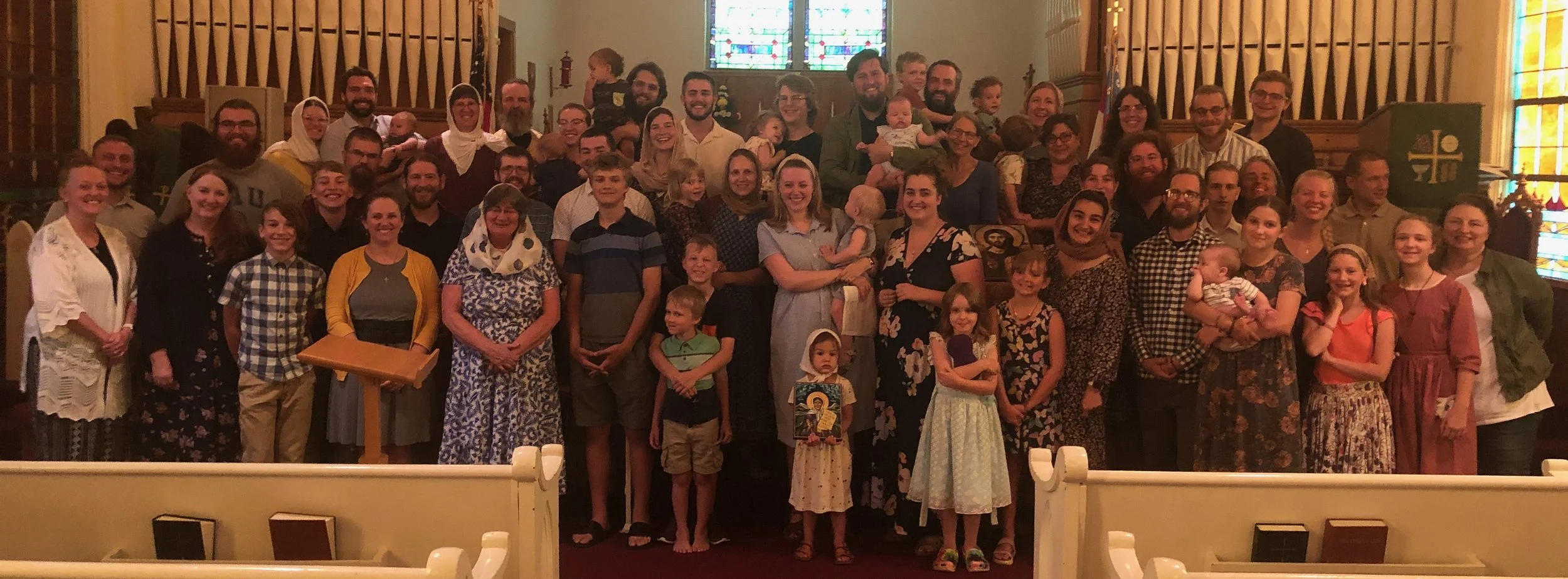 A large group of people gathered inside a church for a photo. The group includes men, women, and children, many of whom are smiling. The church has stained glass windows and a church pulpit at the front.