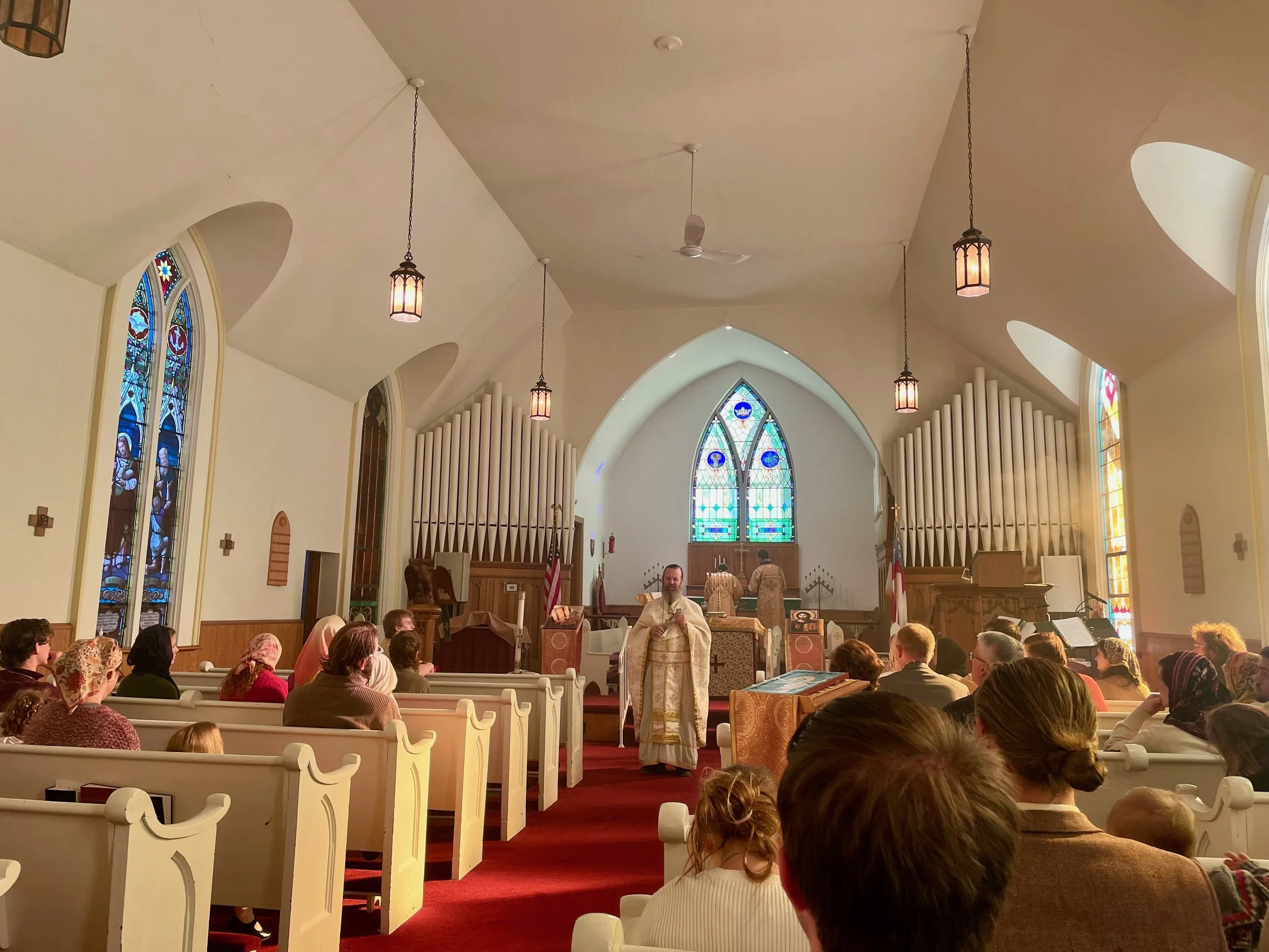 Inside of a church with people attending liturgy, some with head scarves, facing the altar with stained glass windows, organ pipes, and candles at the front.