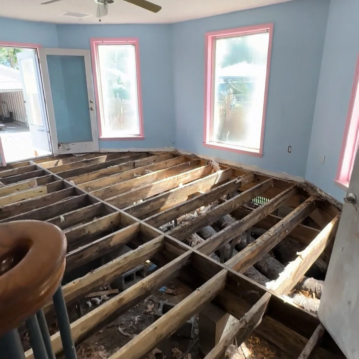 Room under renovation with damaged floorboards removed, exposing the wooden joists and subfloor. Light blue walls with pink window frames and a ceiling fan. Open door leading outside.