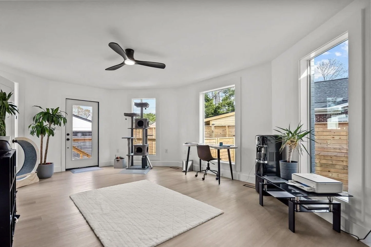 Bright living room with large windows, white walls, light wood flooring, ceiling fan, potted plants, a cat tree, a desk with a chair, and a black TV stand with a printer and a coffee station.