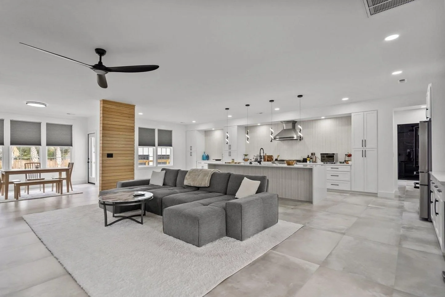 Open-concept living room and kitchen with modern decor, gray sectional sofa, white walls, black ceiling fan, pendant lights over the island, and large windows with gray blinds.