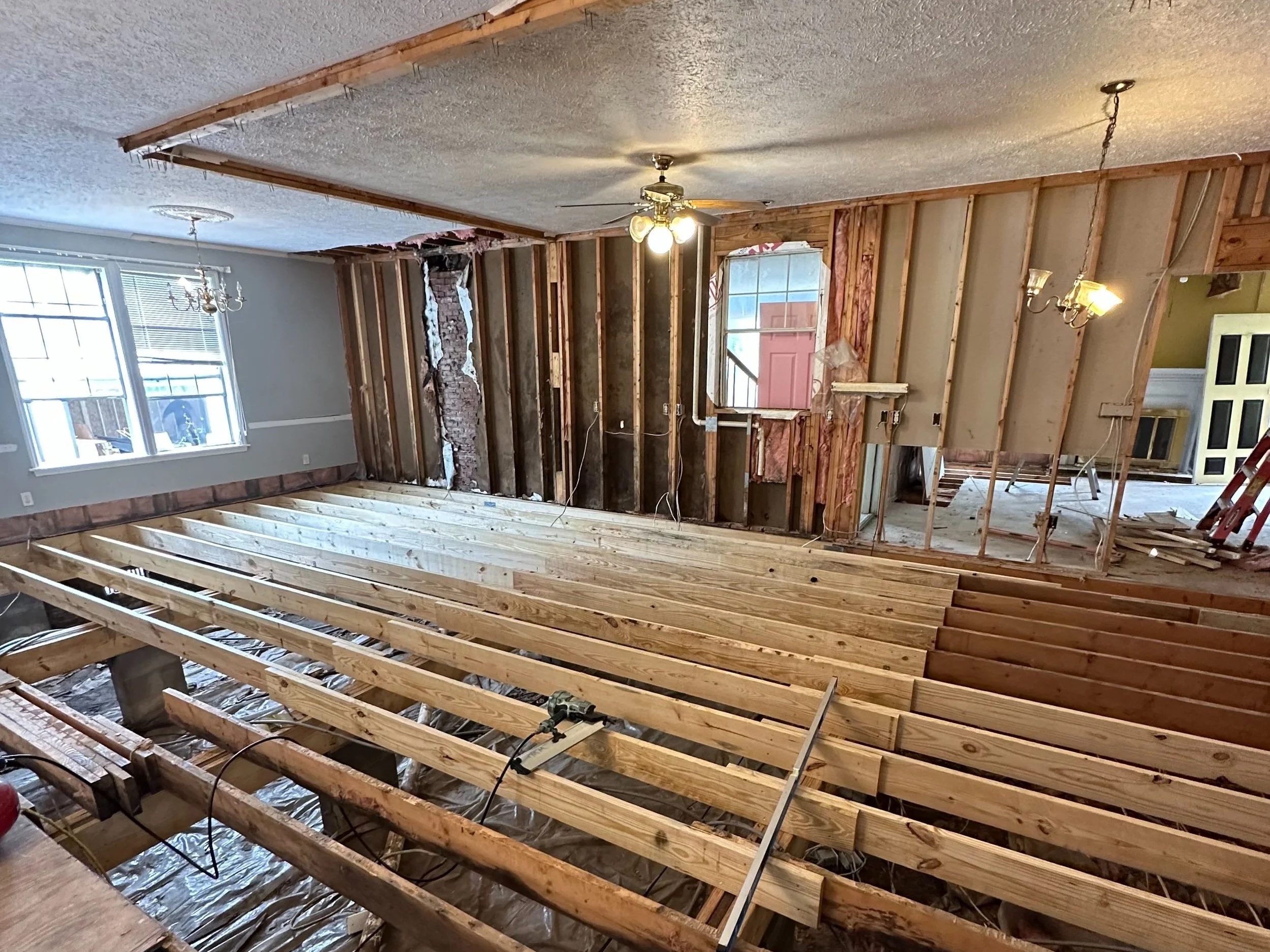 Interior of a house undergoing renovation with exposed wooden floor joists, partially demolished wall, and some light fixtures installed.
