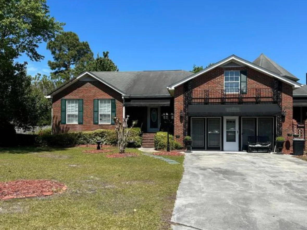A two-story brick house with a driveway leading to a garage, green shutters on the windows, and a small front yard with bushes and trees, under a clear blue sky.