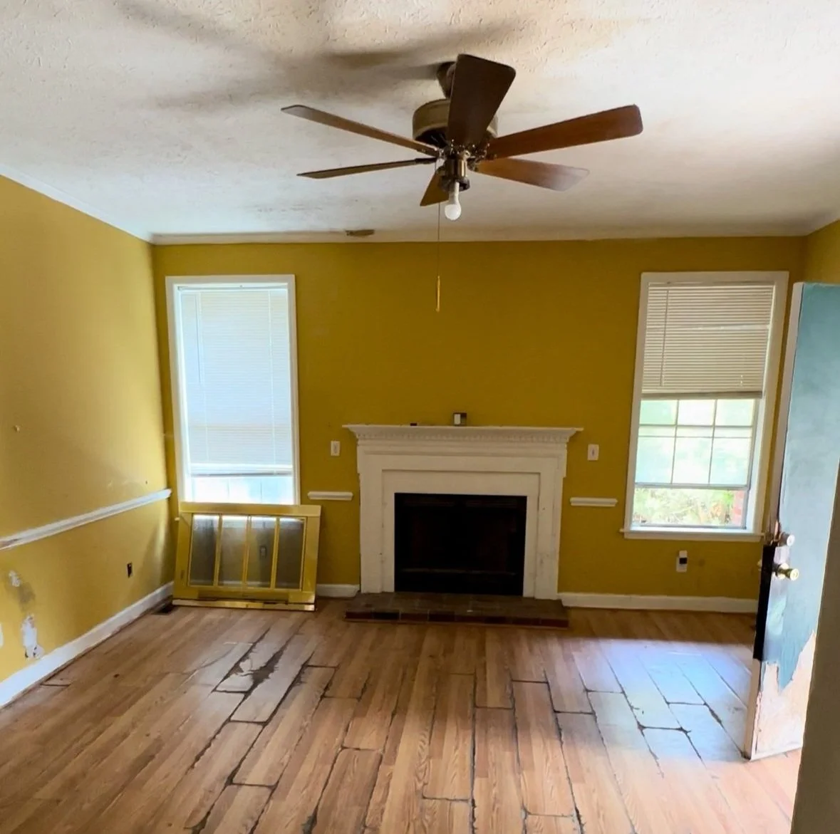 Living room with yellow walls, hardwood floors, a white fireplace, two windows with blinds, a ceiling fan, and a screen on the floor leaning against the wall.
