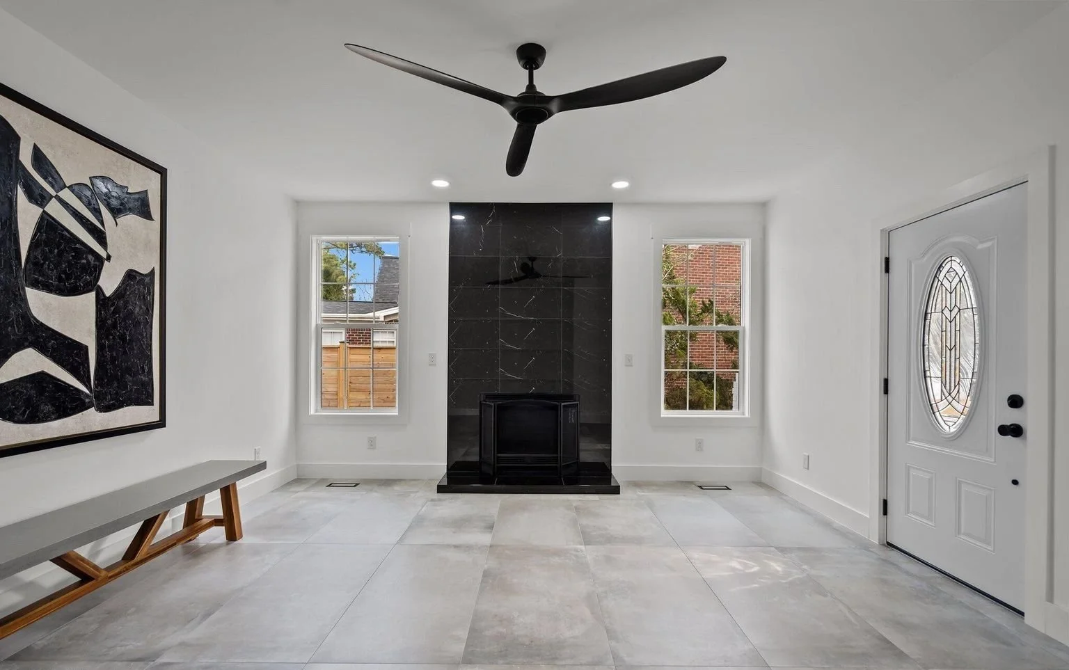 Empty living room with white walls, large tiled floor, black fireplace with marble surround, two windows, a ceiling fan, a front door with oval glass, and a black and white artwork on the wall.