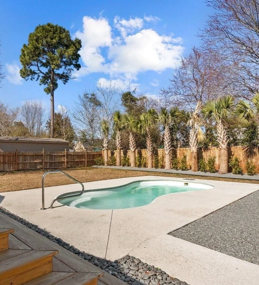 A backyard with a small pool, wooden stairs, and a fence. Palm trees and other trees are in the background under a partly cloudy sky.