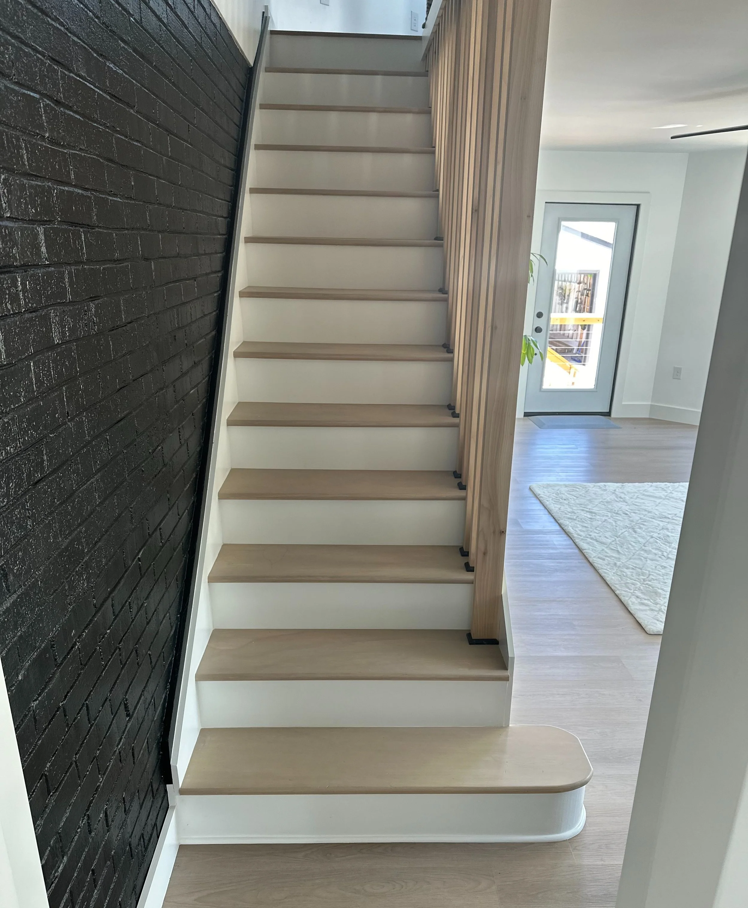 Interior view of a modern staircase with light wood treads, white risers, a black painted brick wall on the left, and a mixed wood and glass door at the top leading outside. The flooring is light wood, and there is a white rug near the door.