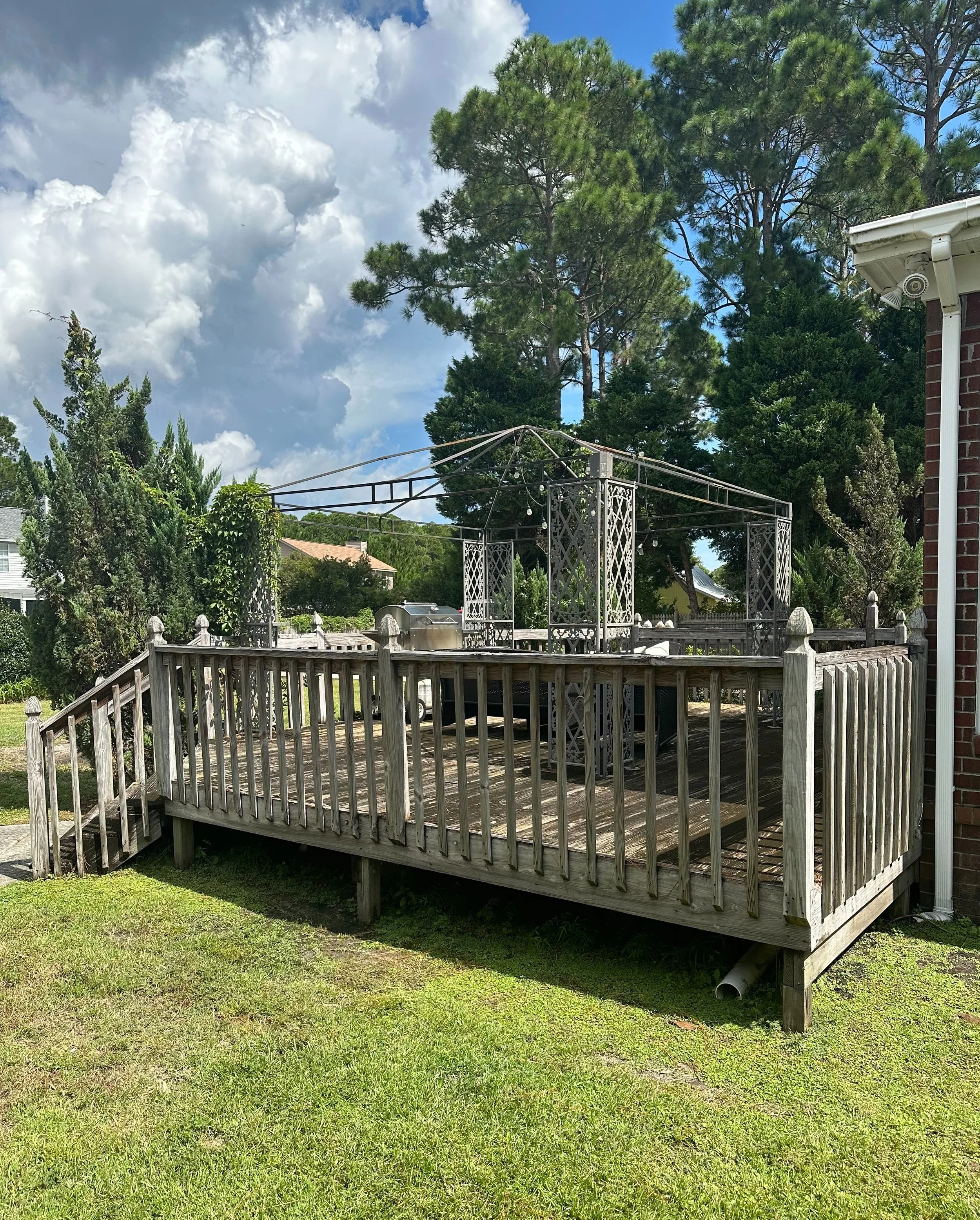 Wooden outdoor deck with a metal frame structure on top, surrounded by trees and a brick wall on the right, with a grassy yard in the foreground and partly cloudy sky overhead.
