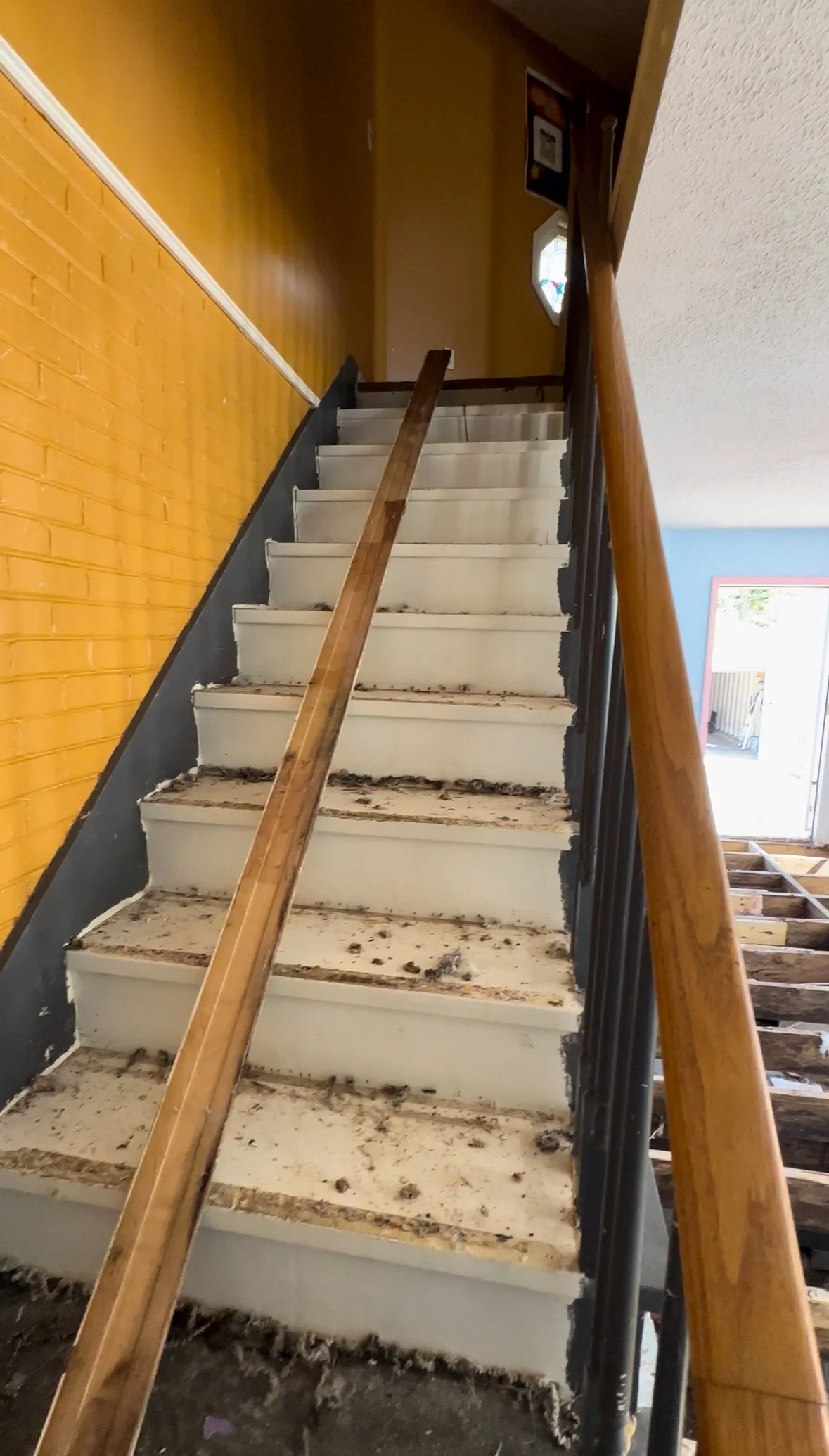 Staircase under renovation with missing and dirty steps, black railing on the right, yellow wall on the left, and a wooden handrail.