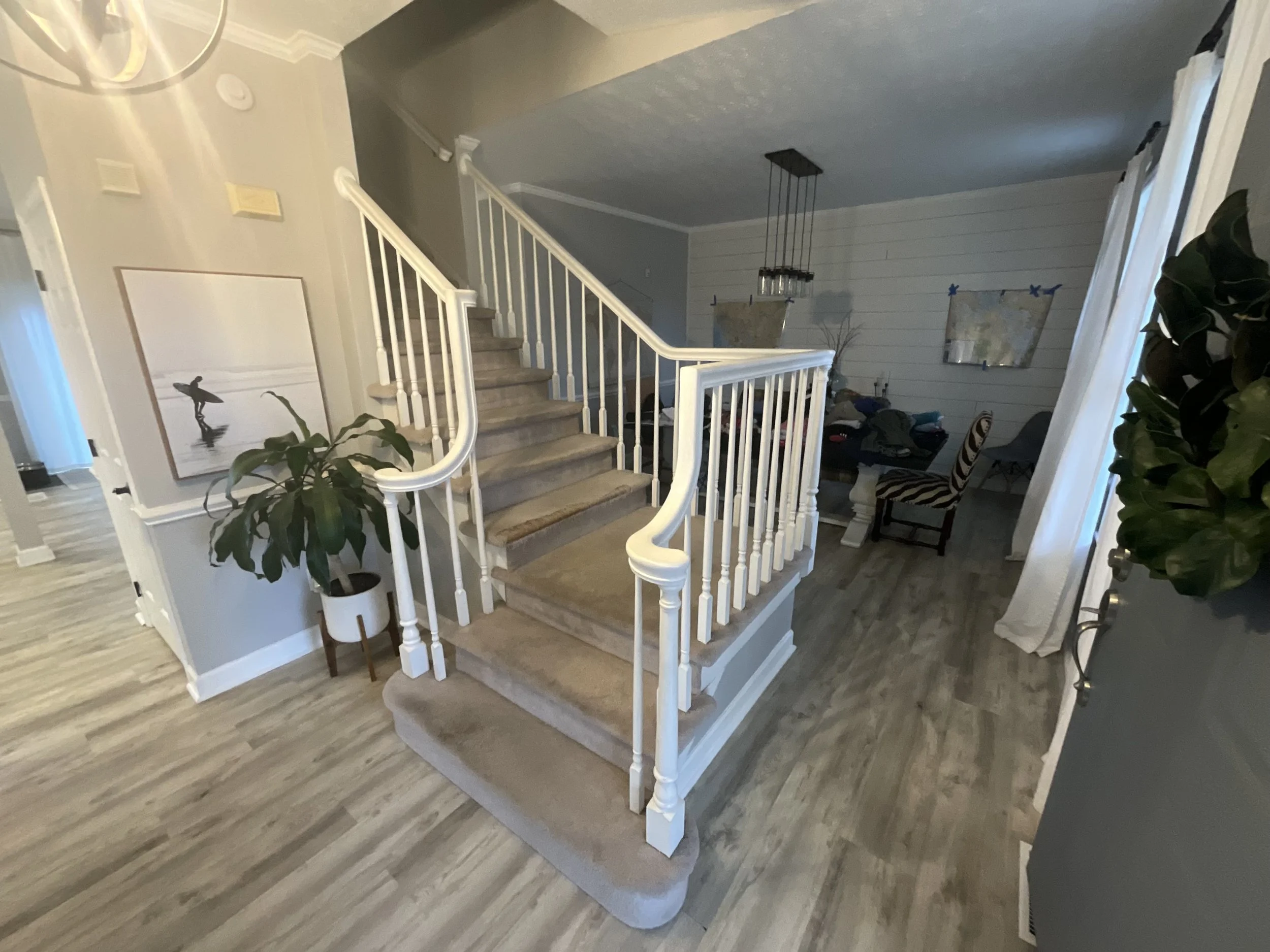 Interior view of a home featuring a staircase with white railings and beige carpet, a potted plant beside the stairs, and a dining area with a table and chairs in the background, with white curtains on windows.