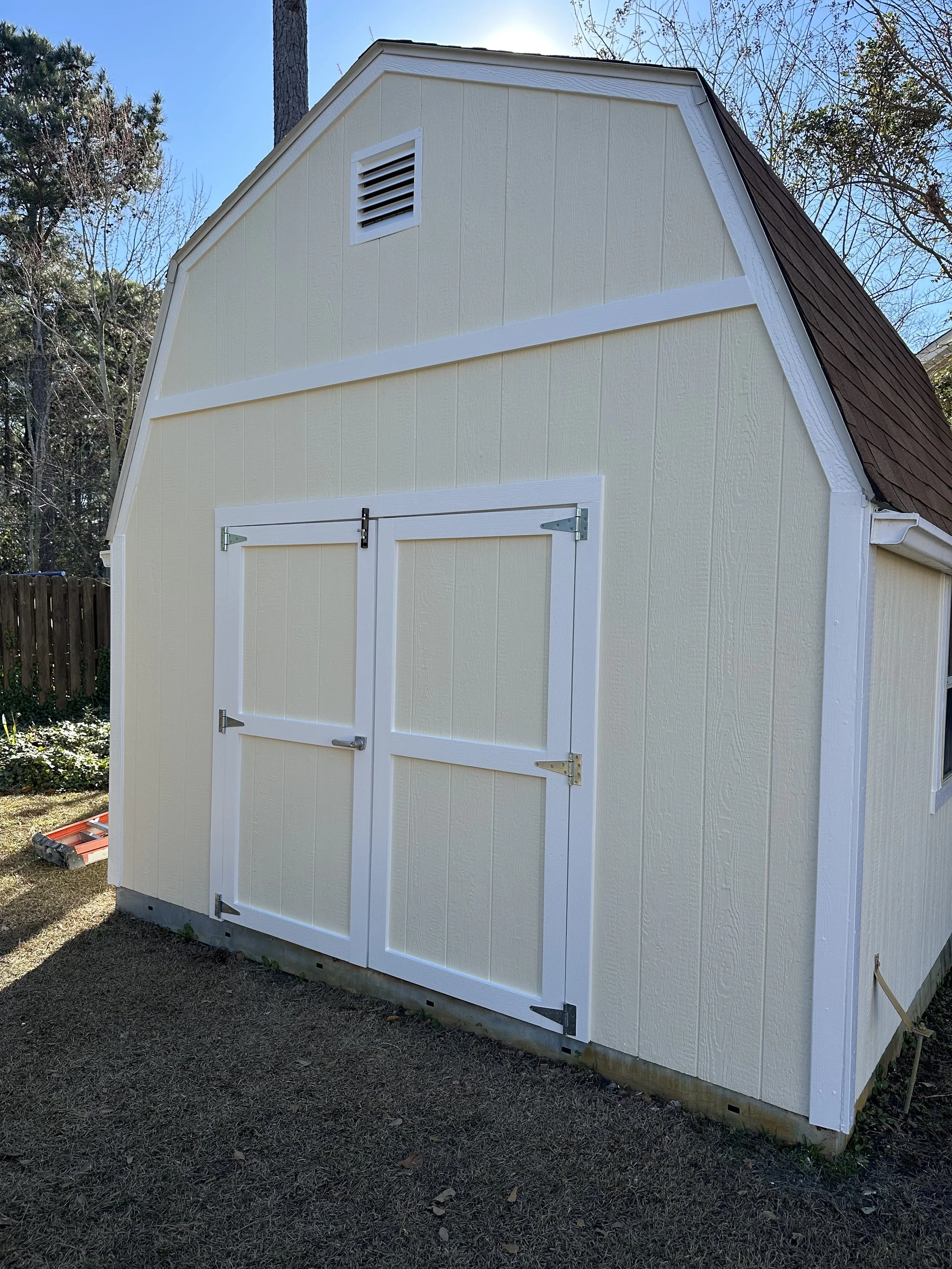 A small, cream-colored shed with white trim and double doors, background of trees and a clear blue sky.
