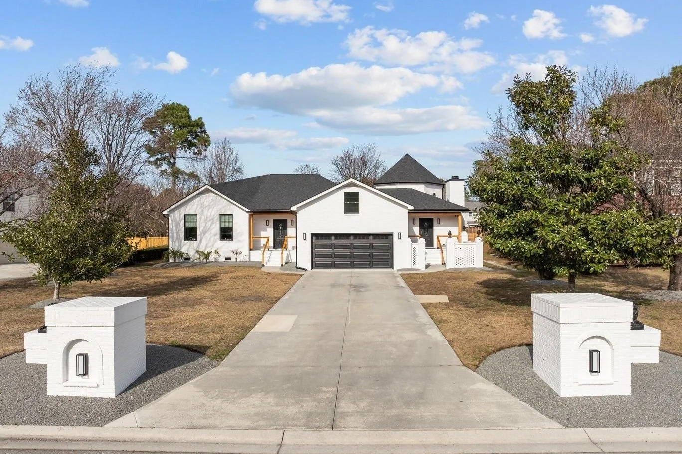 Modern white house with a black garage door, front driveway, and landscaped yard with trees, and decorative white brick walls with lanterns on posts.