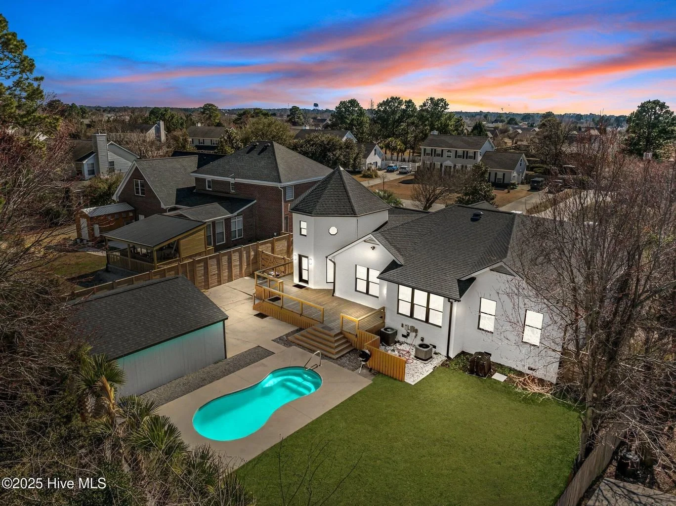 Aerial view of a suburban neighborhood at sunset, featuring a house with a backyard pool, a wooden deck, and fenced yard, surrounded by similar houses and trees.