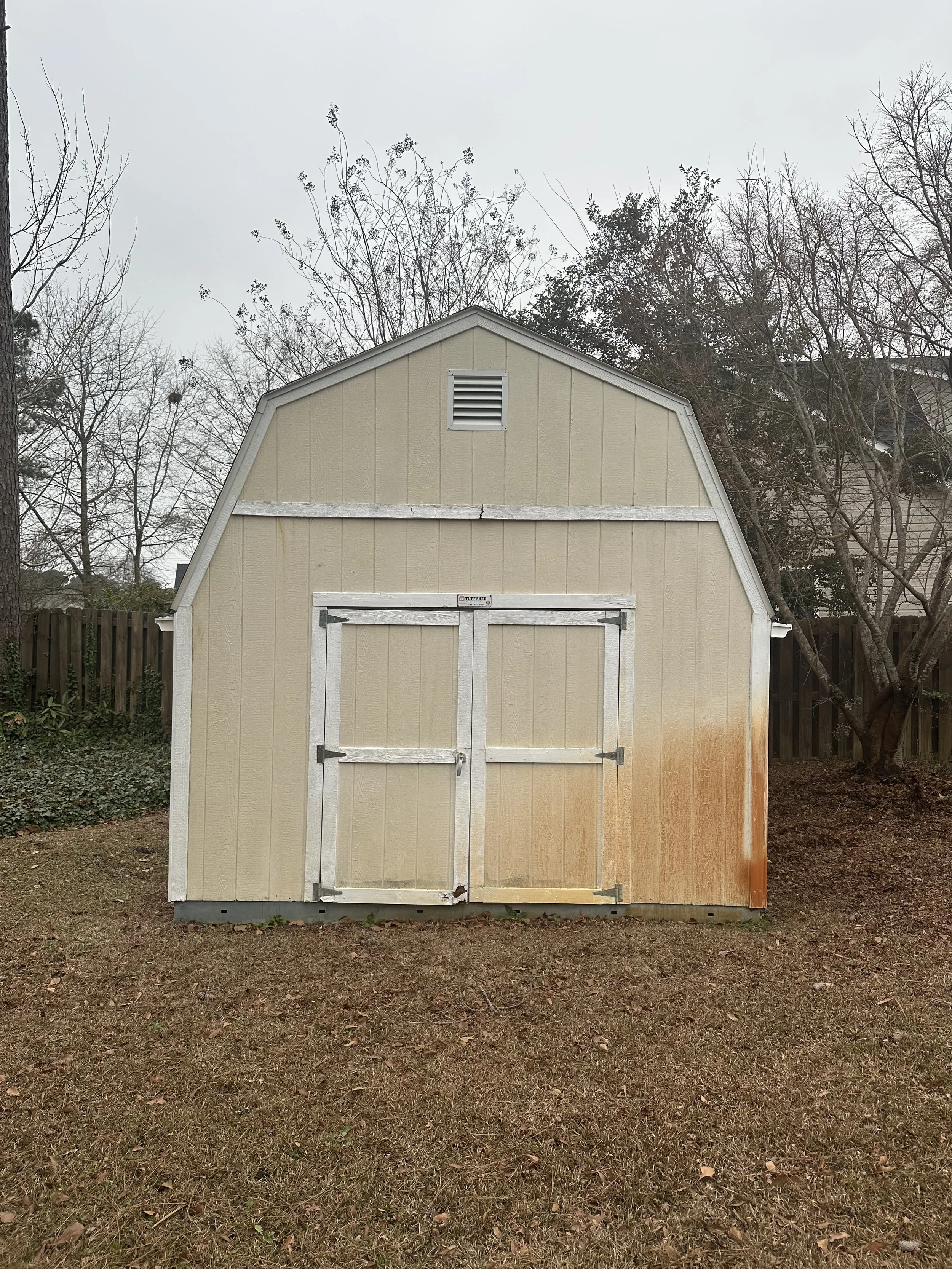 A beige shed with white trim, double doors, and a vent near the roof, located in a backyard with leafless trees and a wooden fence, on an overcast day.
