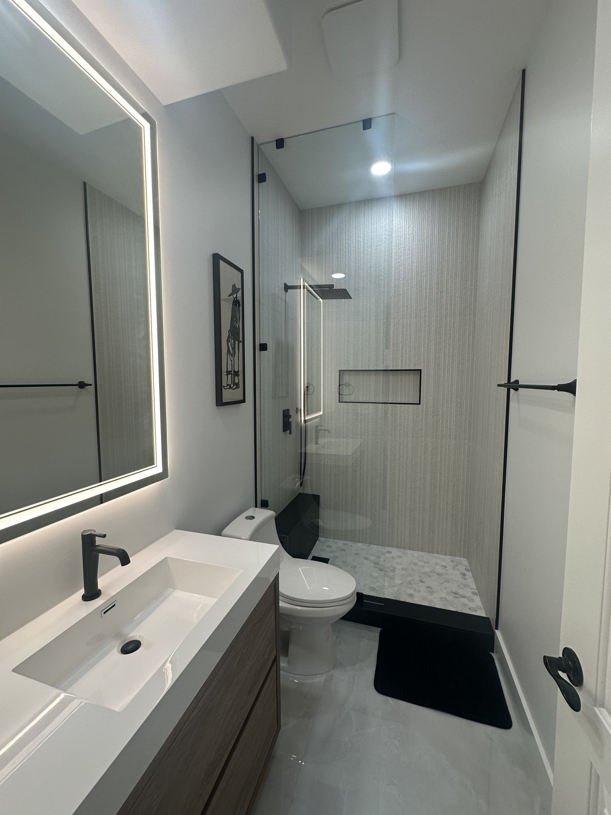 Modern bathroom with white sink, black fixtures, large mirror with lights, toilet, glass shower with built-in niche, black shower fixtures, black towel bars, and a black bath mat.