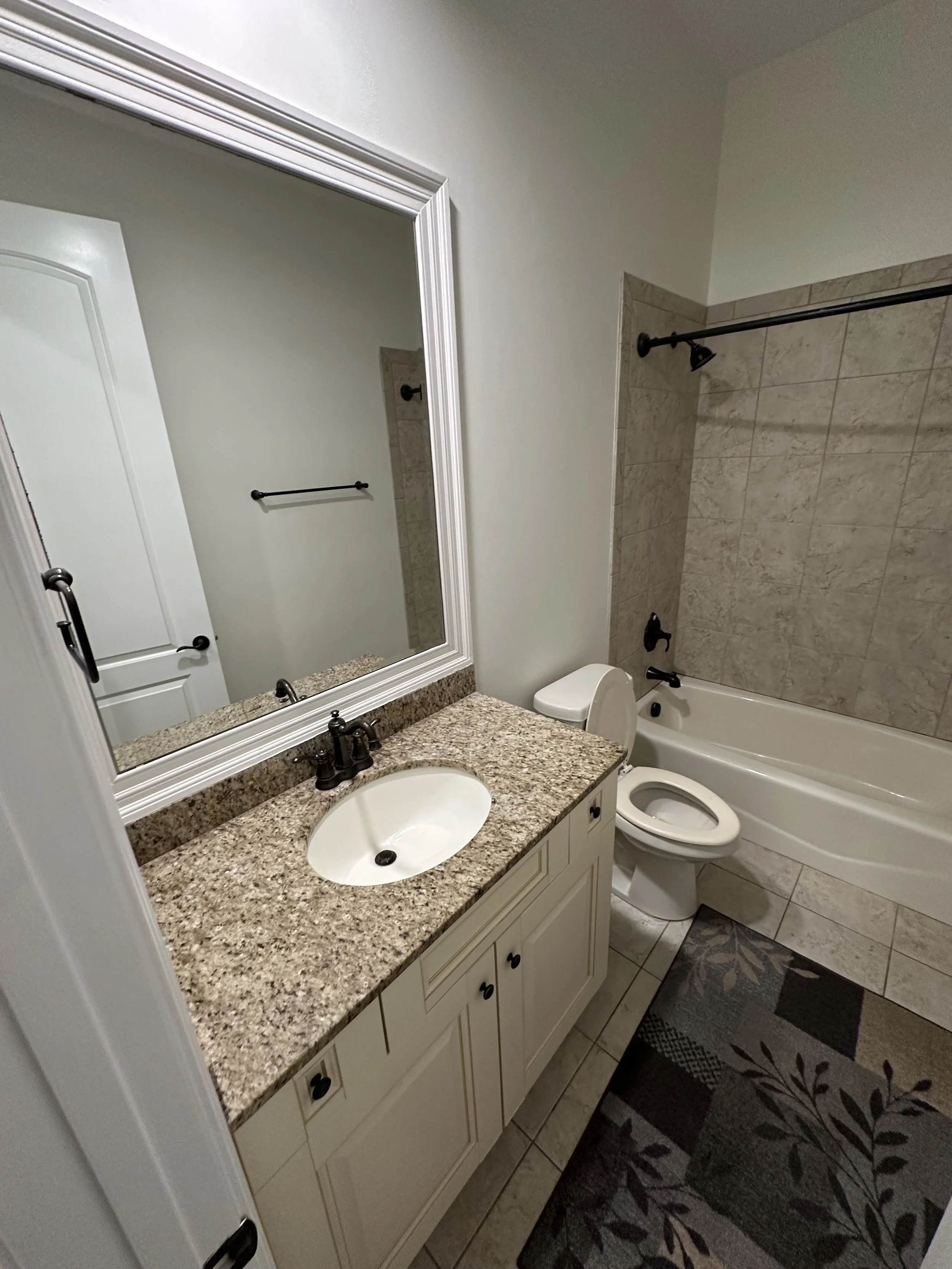 Bathroom with a granite countertop sink, large mirror, bathtub with beige tiles, toilet, and patterned rug on floor.