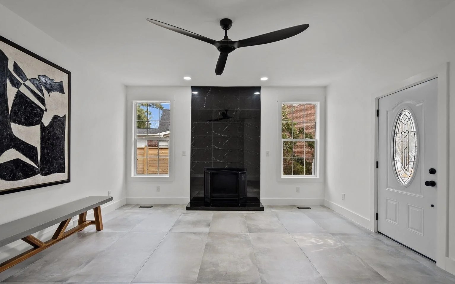 Empty modern living room with white walls, large tiled floor, black fireplace with marble tiles, two windows, a ceiling fan, and a white door with oval glass window.