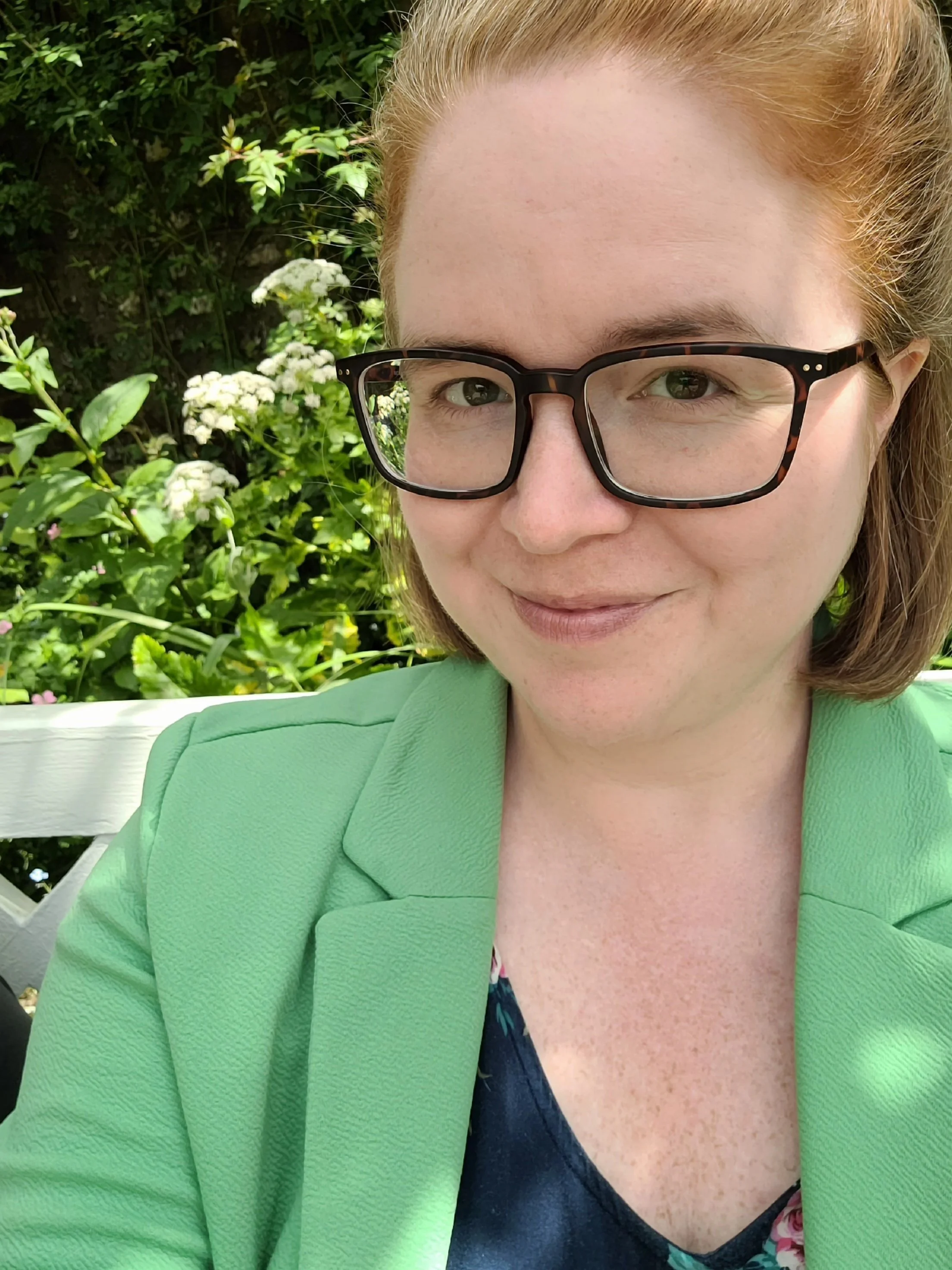 A woman with red hair, wearing glasses and a bright green blazer, taking a selfie outdoors with greenery and white flowers in the background.