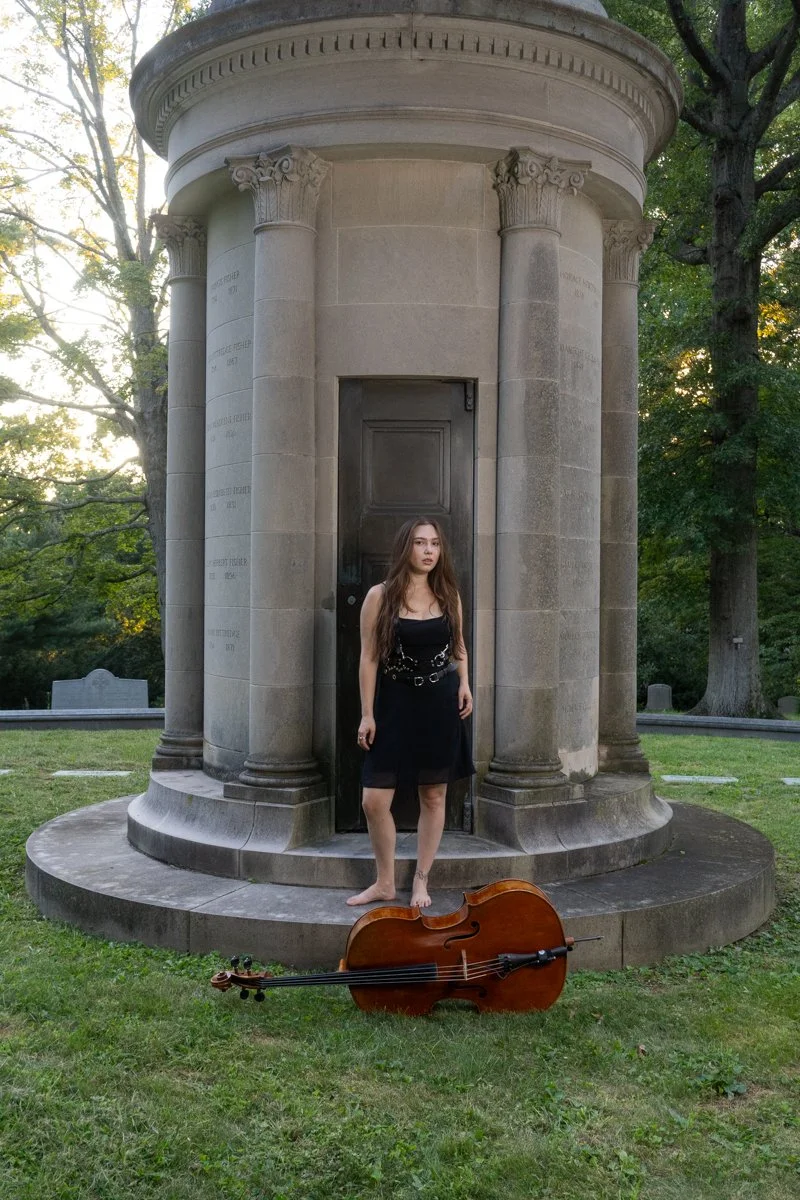 A young woman in a black dress standing barefoot on the steps of a stone monument, with a cello lying on the grass in front of her, in a cemetery with tall trees and green foliage.