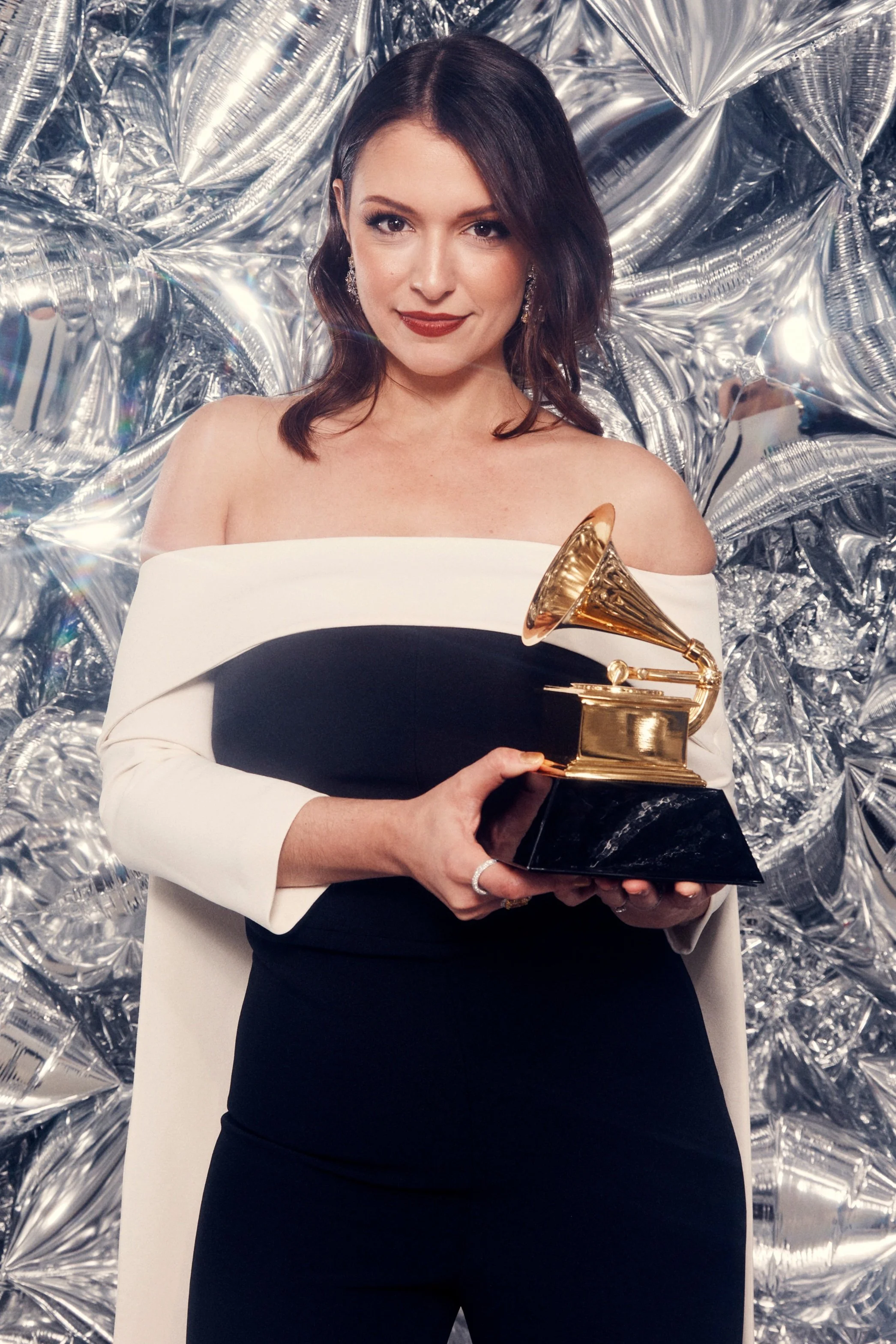 Woman in formal attire holding a Grammy Award trophy, standing in front of a metallic, reflective backdrop.