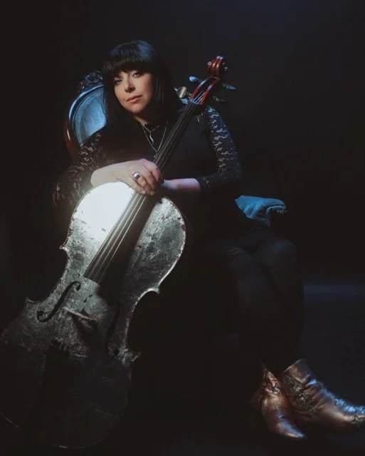 A woman sitting in a dark setting holding a large, worn acoustic cello, wearing a black lace top and brown boots.
