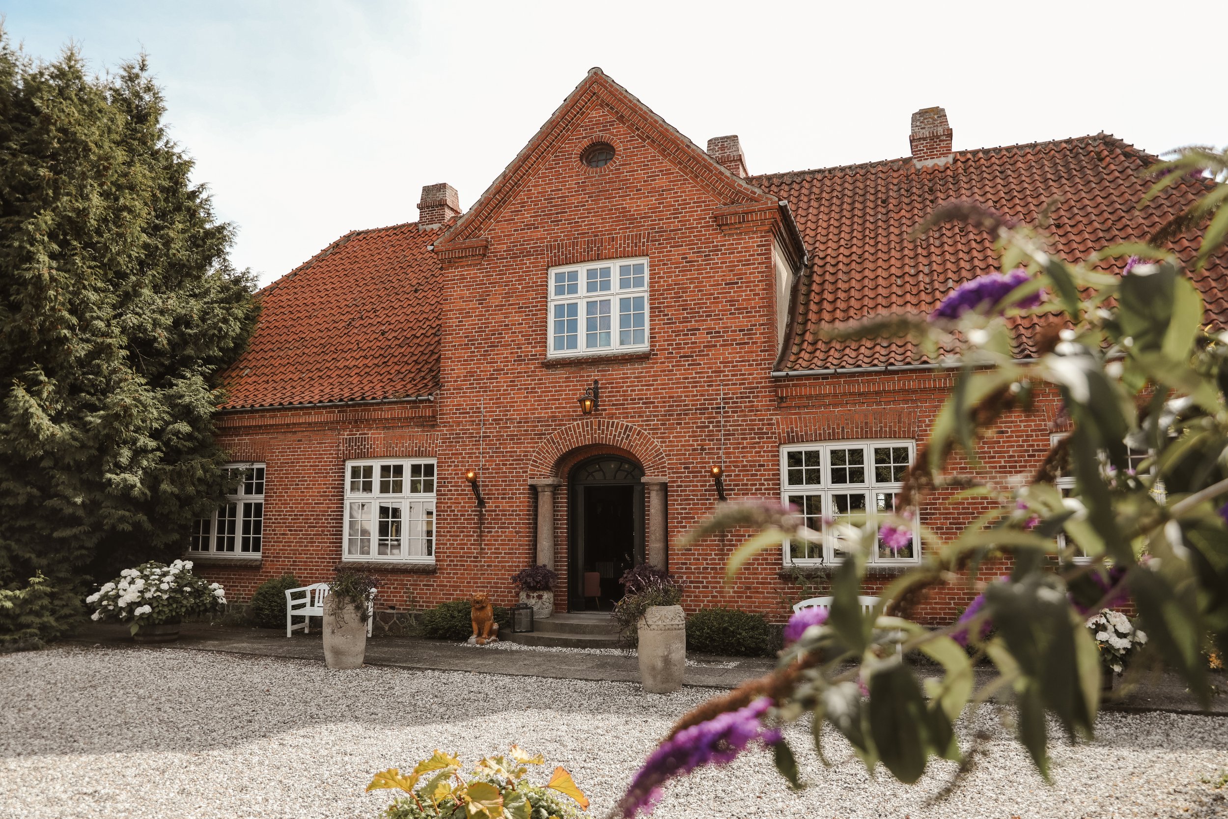 Tegelhus i traditionell stil med rött tak, omgiven av grus och blommor i förgrunden.