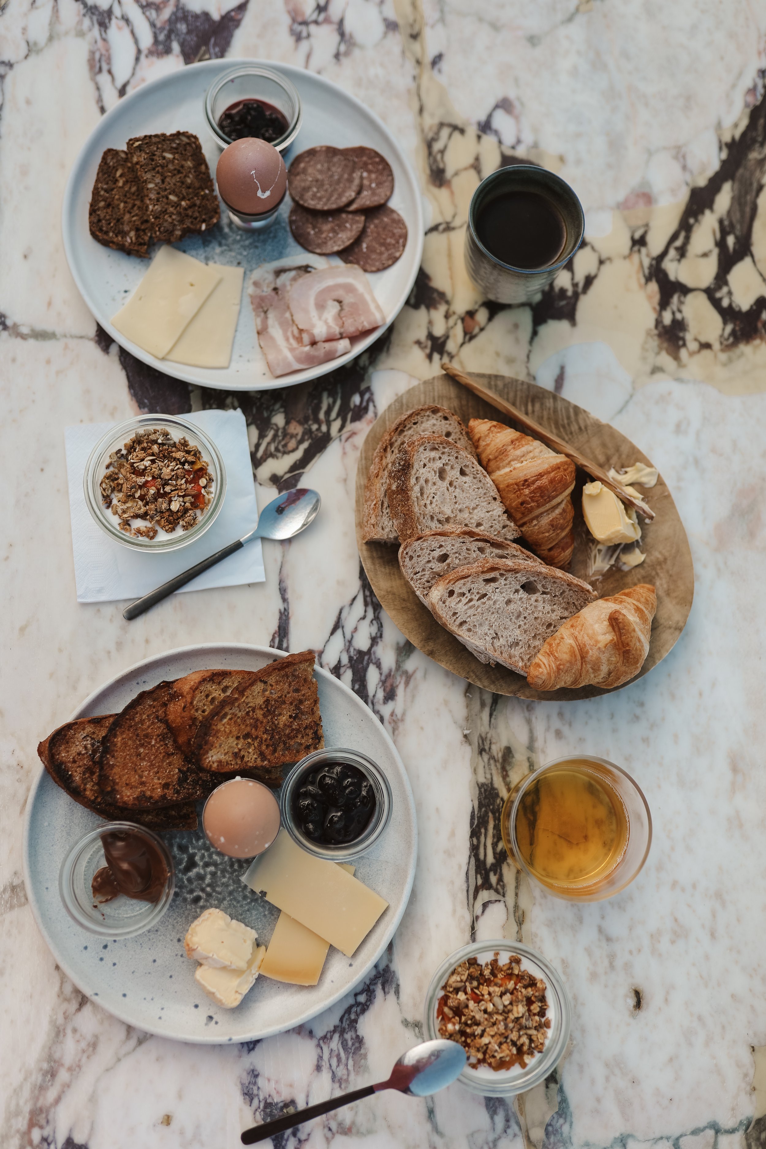 Frukost på ett marmorbord med bröd, ost, ägg, charkuterier, yoghurt med müsli, smörcroissanter och drycker.
