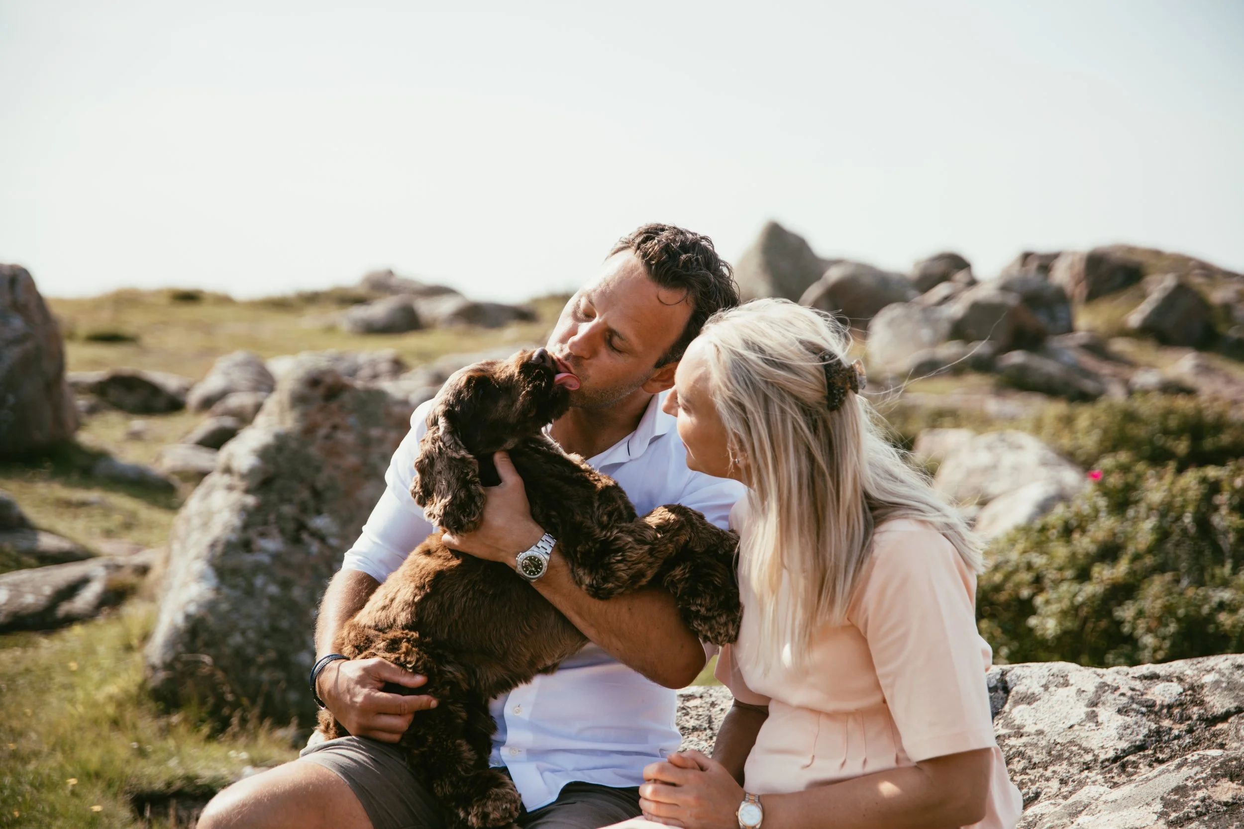 Två människor sitter utomhus på stenar med en brun hund i famnen. Mannen pussar hunden medan kvinnan tittar på."}