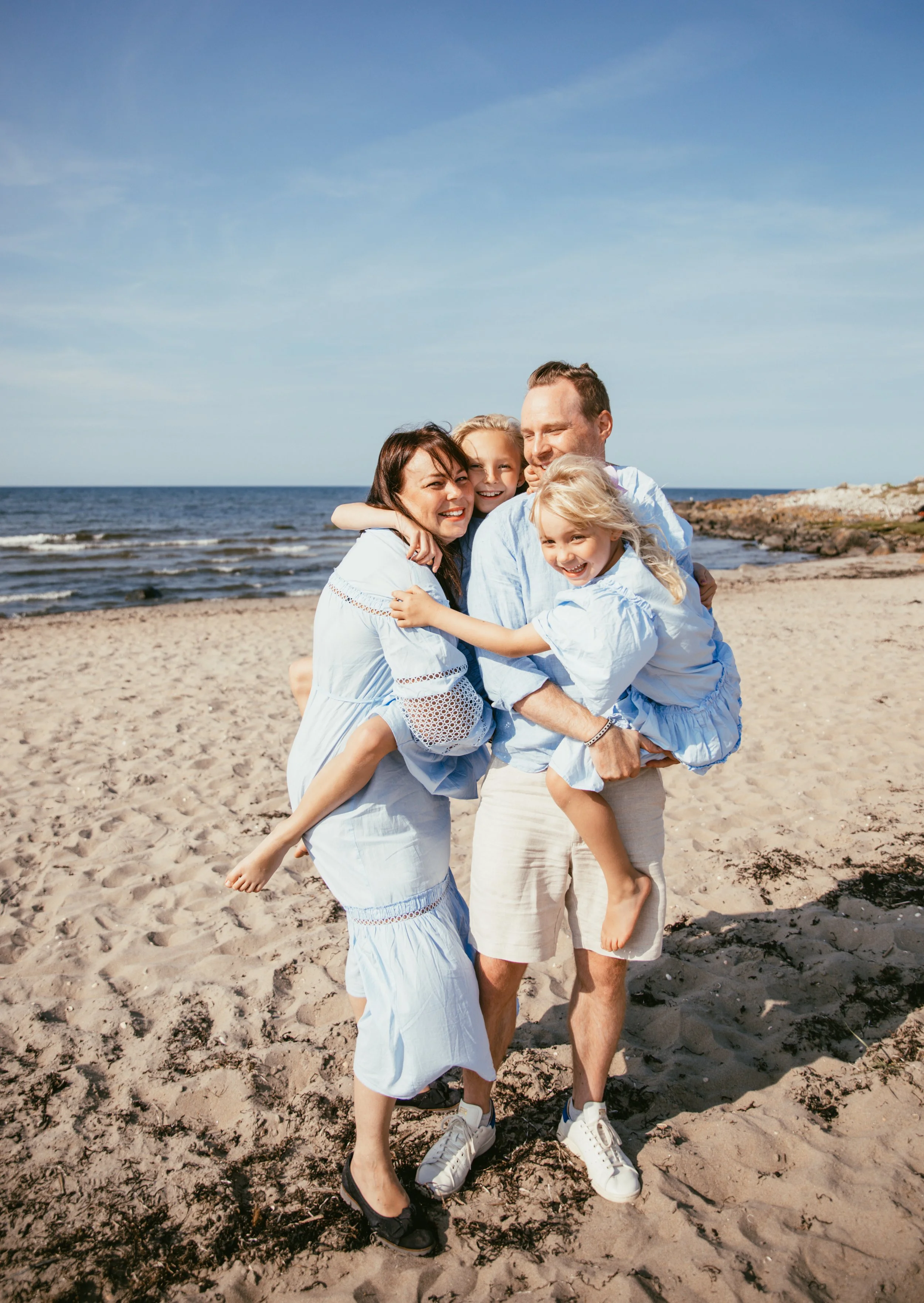 En familj med två vuxna och två barn kramas på en strand. Alla är klädda i ljusblå kläder och verkar glada. Havet och molnfri himmel syns i bakgrunden.