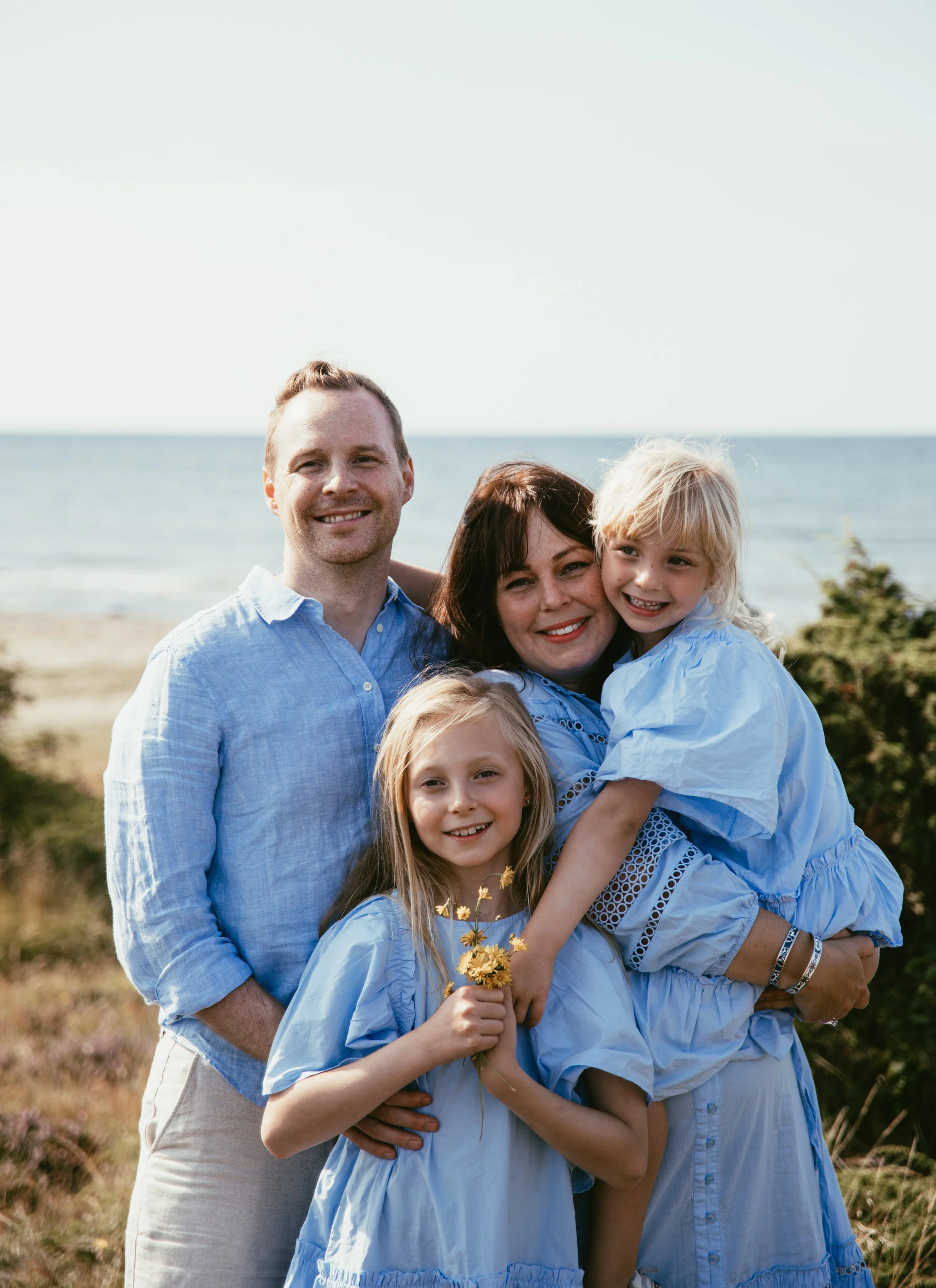 En familj står tillsammans vid havet, alla klädda i ljusblå kläder. Två barn är framför föräldrarna, medan en vuxen håller en bukett av gula blommor.