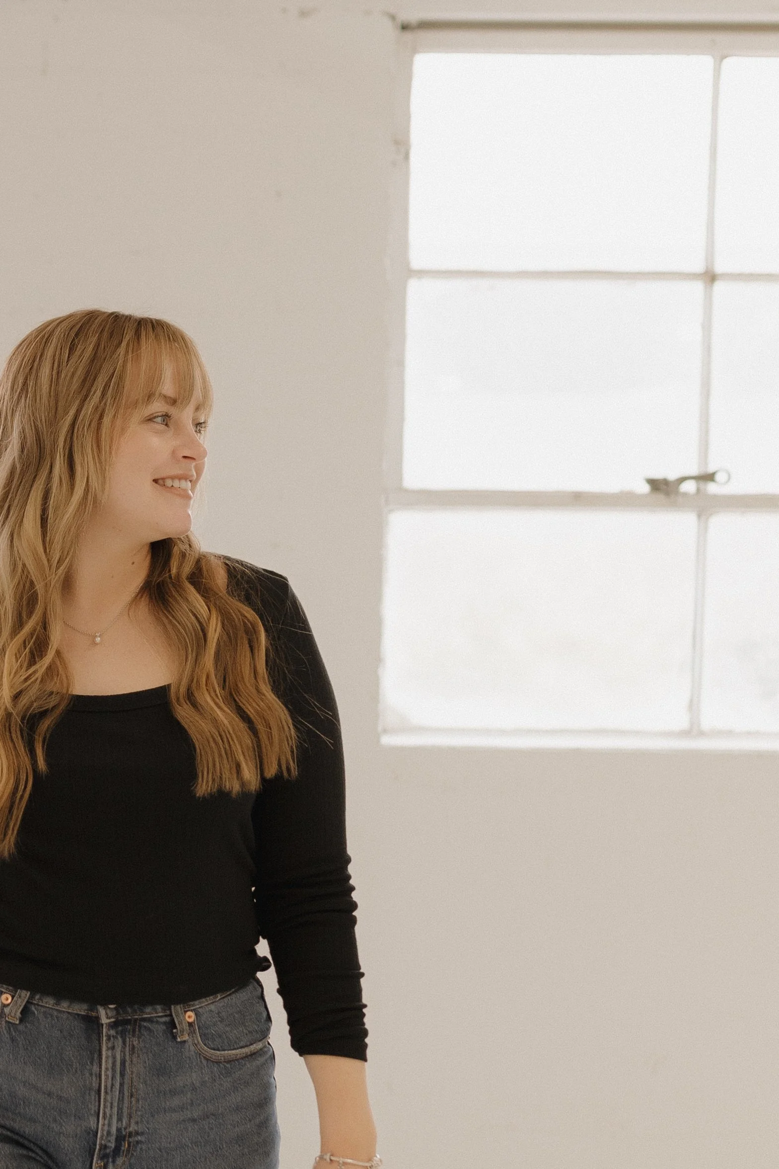 A young woman with long, wavy, blonde hair, smiling and looking to her left, standing indoors near a window with a metal latch and a plain wall. Elopement planner for Western Slope of Colorado.