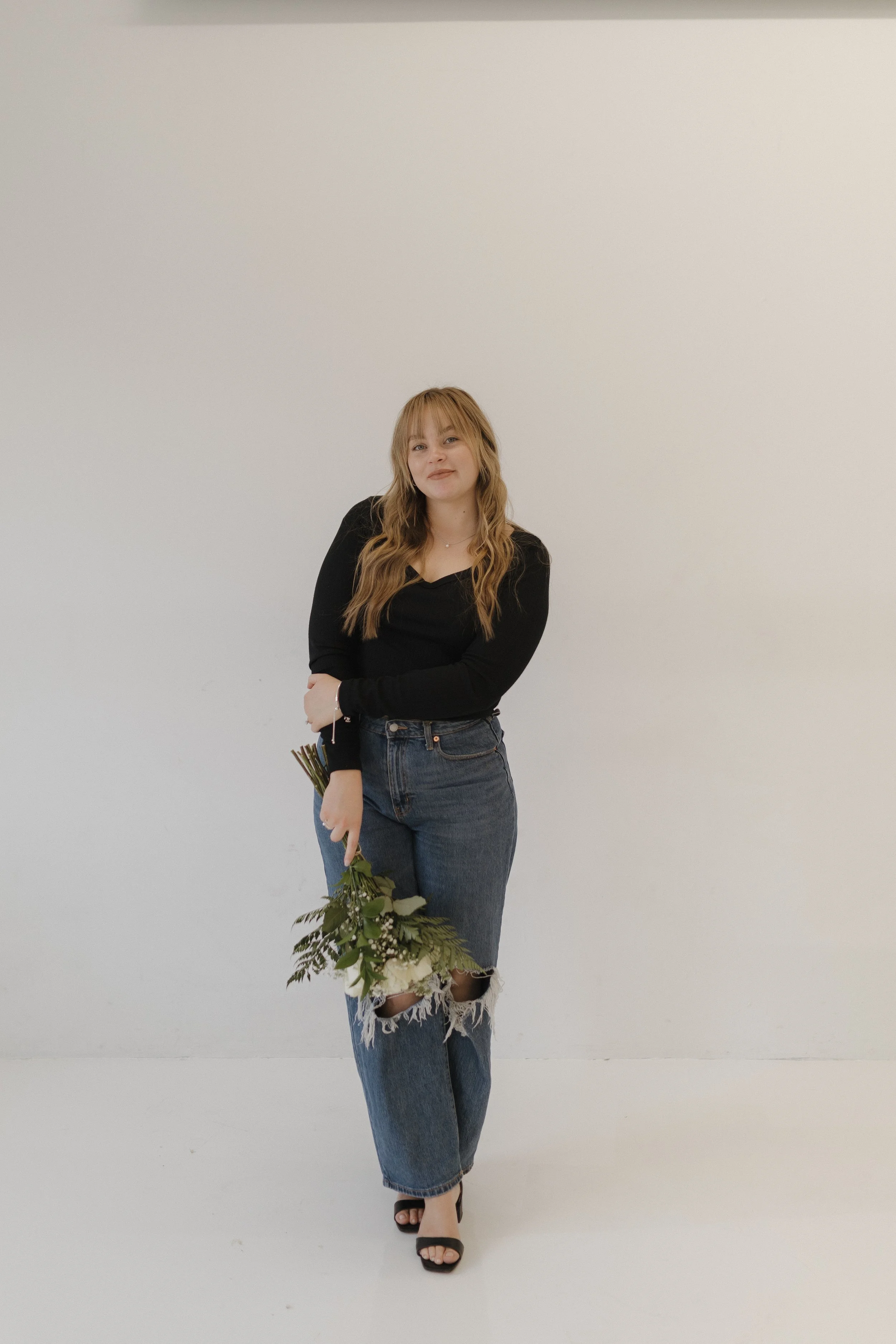 A young woman with long wavy blonde hair stands against a plain white wall, holding a bouquet of flowers, wearing a black long-sleeve top, ripped jeans, and black heeled sandals. Micro-wedding planner in Western Colorado by Timeless Traditions