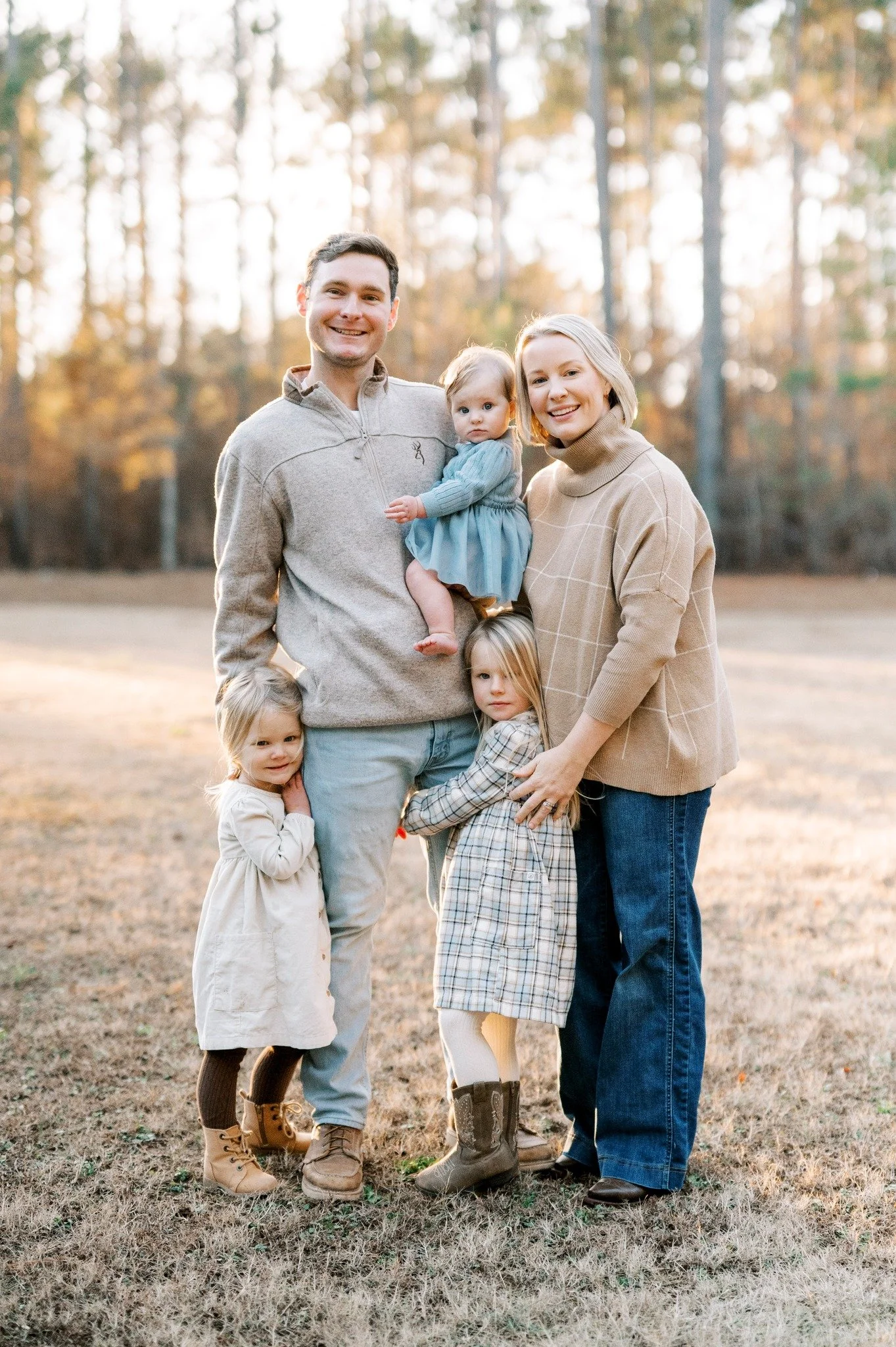 A family of five standing outdoors on a grassy field during autumn, with tall trees in the background. The father, mother, and three young daughters are smiling and posing for the photo.