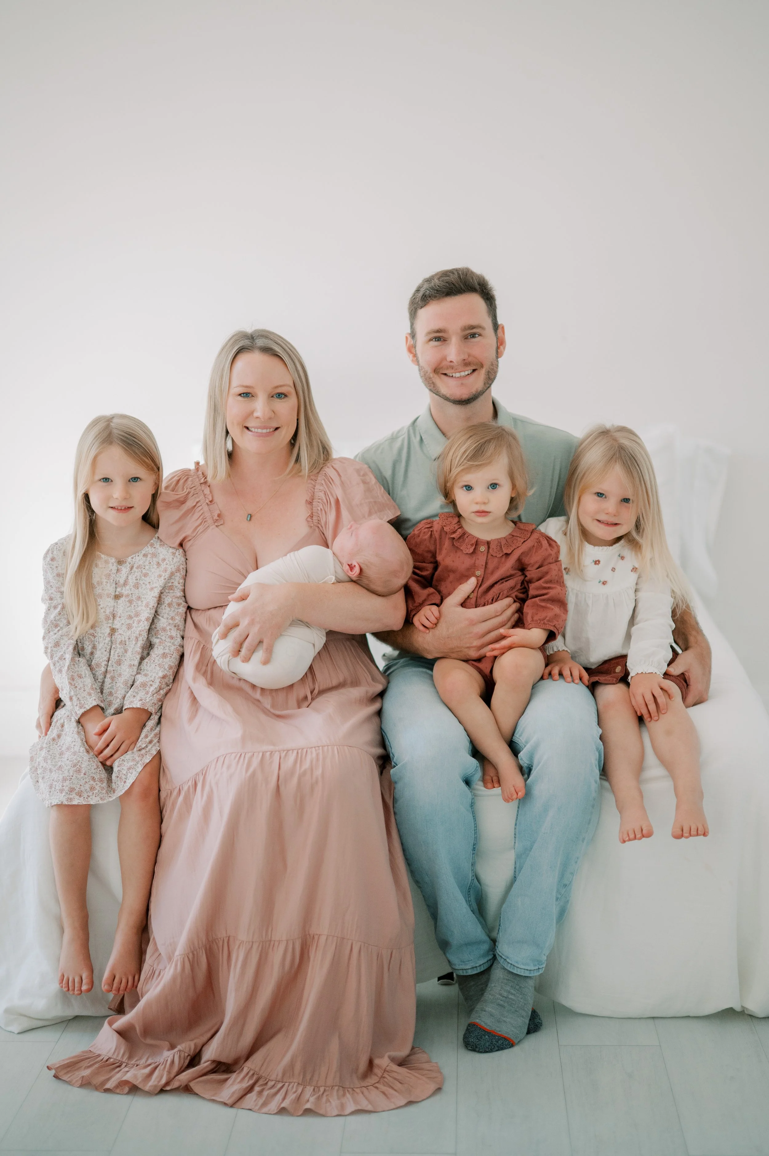 A family of five sitting on a white couch, with a woman holding a newborn baby, two young girls, and a man. They are smiling and dressed in casual clothing in a bright, minimalistic room.