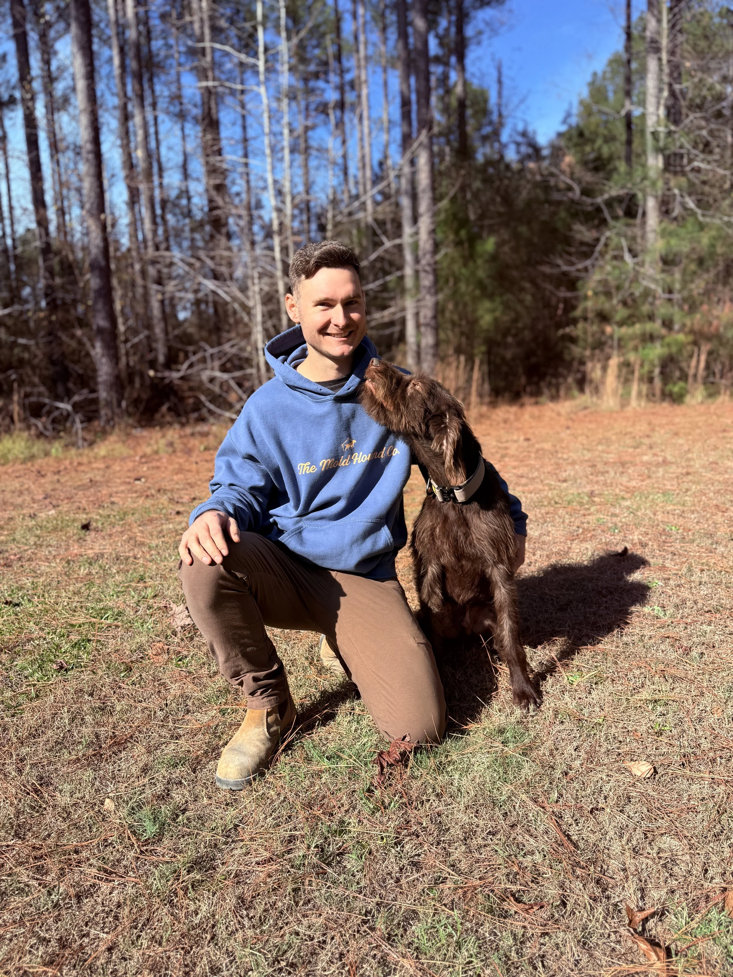 A man kneeling on grass in a forested area, wearing a blue hoodie and brown pants, with a brown dog next to him, and the dog licking his face.