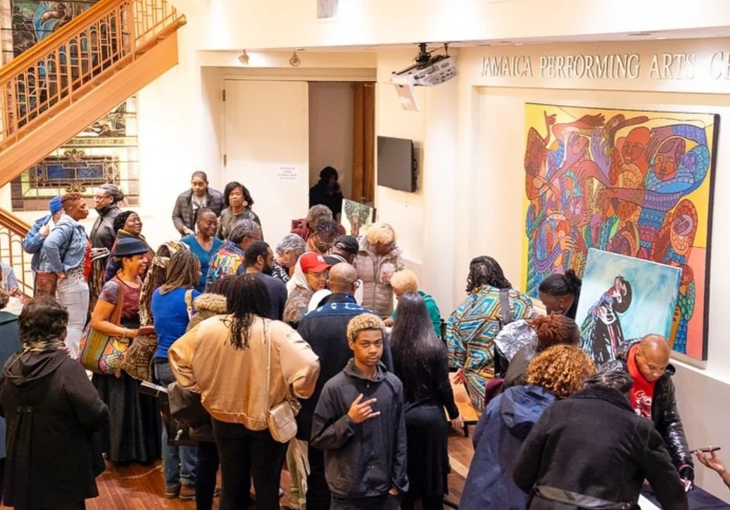 Crowd of people gathered in an art gallery with colorful paintings on the wall, a staircase to the left, and a sign that reads 'Jamaica Performing Arts Center'.