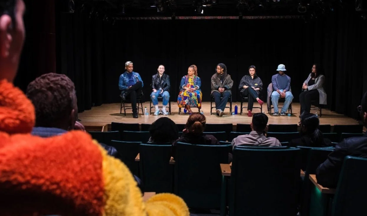 Panel discussion with seven women seated on stage in front of black curtains, facing an audience in a theater or auditorium.