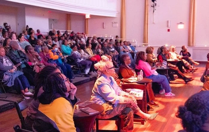 Audience seated and attentively listening at an indoor event, with some people taking notes, in a well-lit auditorium.