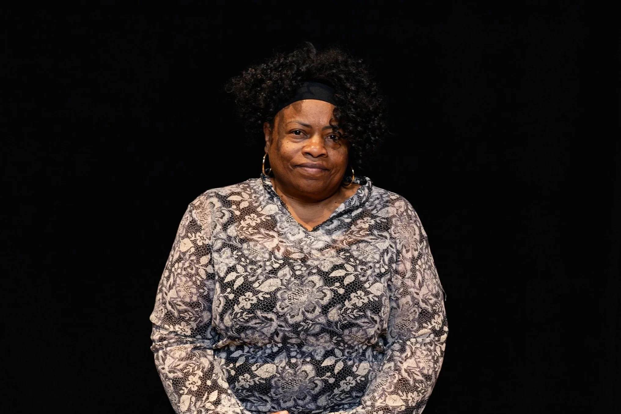 Close-up of a smiling woman with dark curly hair outdoors in a grassy area with trees in the background.