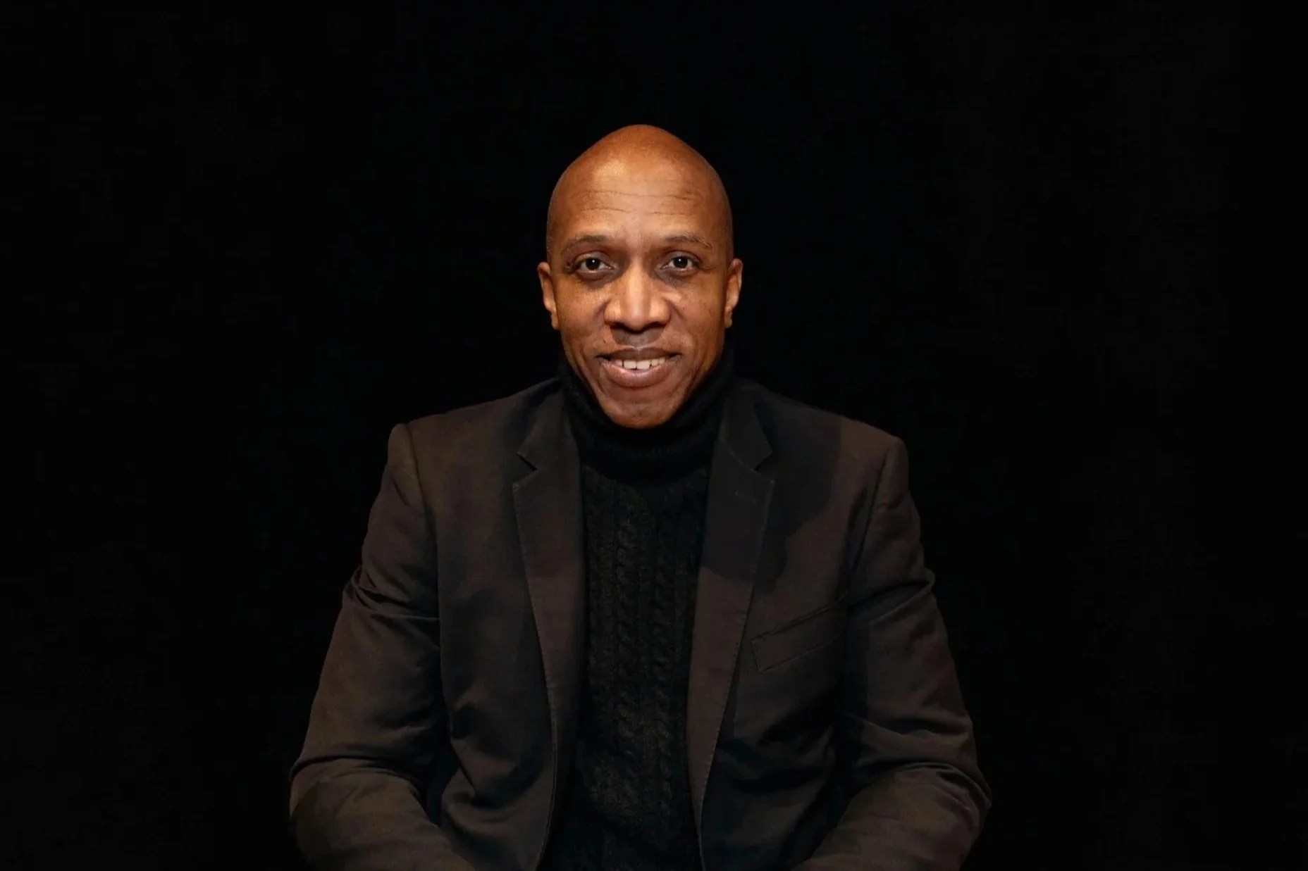 Man in a suit with hands clasped, wearing a watch, looking into the camera.