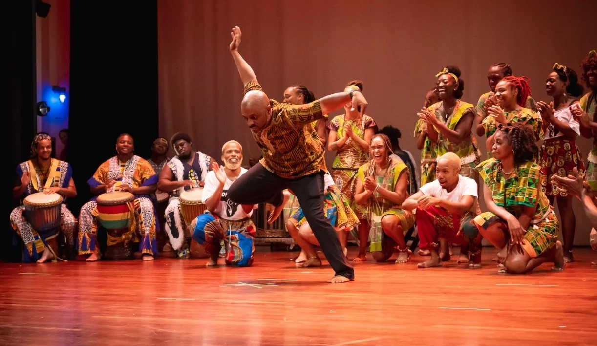 A group of people performing traditional African dance and drumming on stage, smiling and clapping.