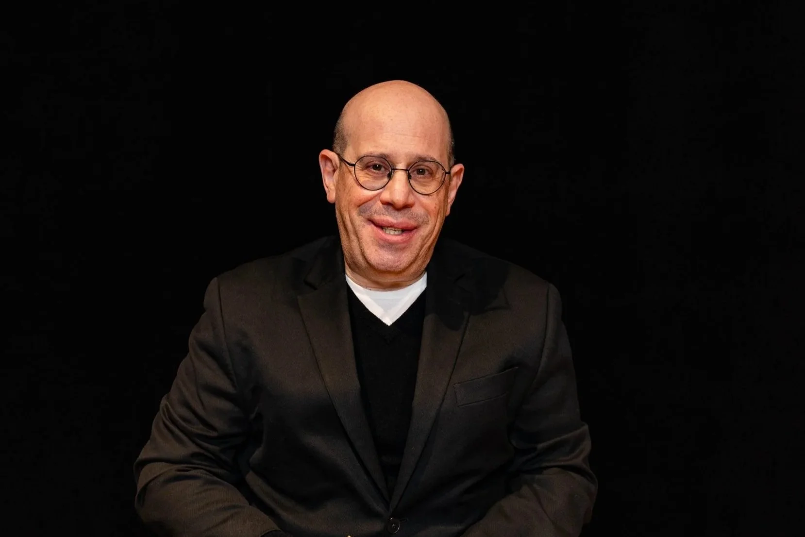 Close-up headshot of a bald man with light skin, wearing a blue shirt and navy blazer, against a black background.
