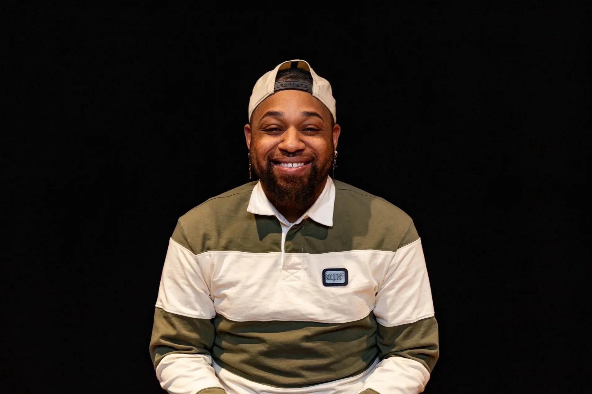 A man smiling outdoors in front of a brick building wearing a leather jacket and hat.