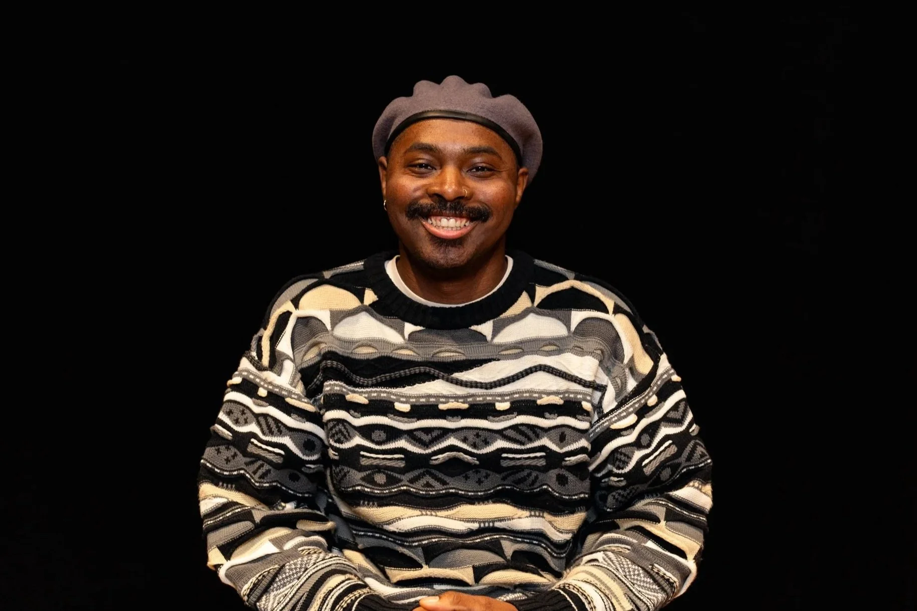 A man in a brown leather jacket and brown beanie leaning against a pole on a city street at night, with a blurred background of construction scaffolding and lights.